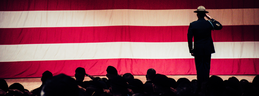 Silhouette of a military officer saluting in front of an American flag with an audience in shadow.