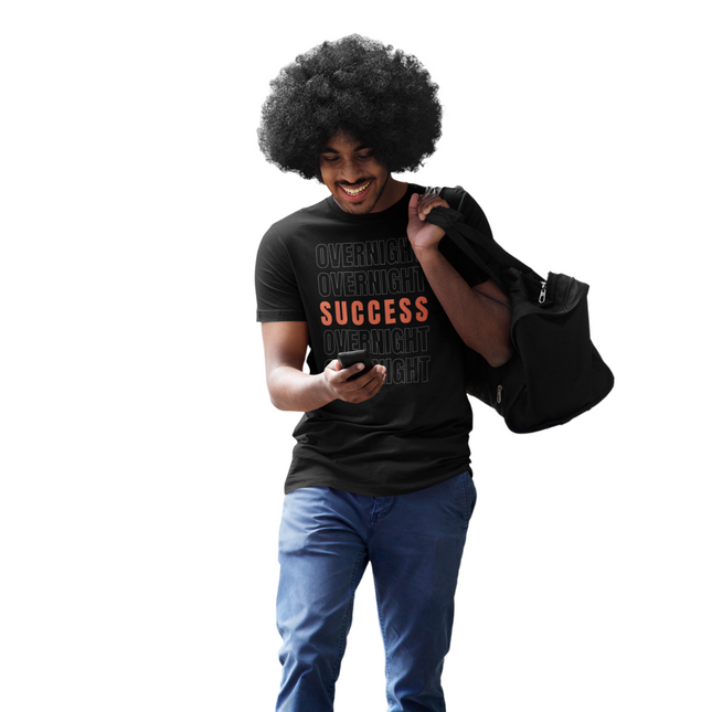 Overnight Success Is a Myth T-Shirt – Built for the Hustlers worn by a man with a bag, smiling and using a phone.