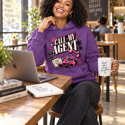 A woman sitting at a wooden table in a bright cafe setting, wearing a purple hoodie with a graphic that reads 'CALL MY AGENT' and holding a white mug that says 'Good things come from everywhere.'.