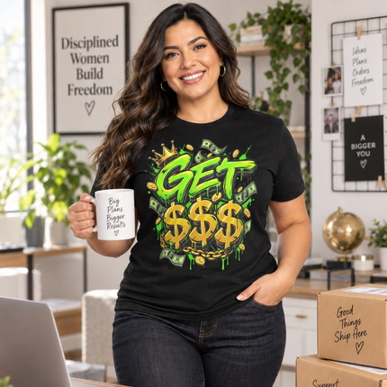 A woman stands in a bright home office wearing a black graphic T-shirt with a green and gold 'GET $$$' design. She holds a white mug and has a laptop, phone, and cardboard shipping boxes on a wooden desk in front of her.