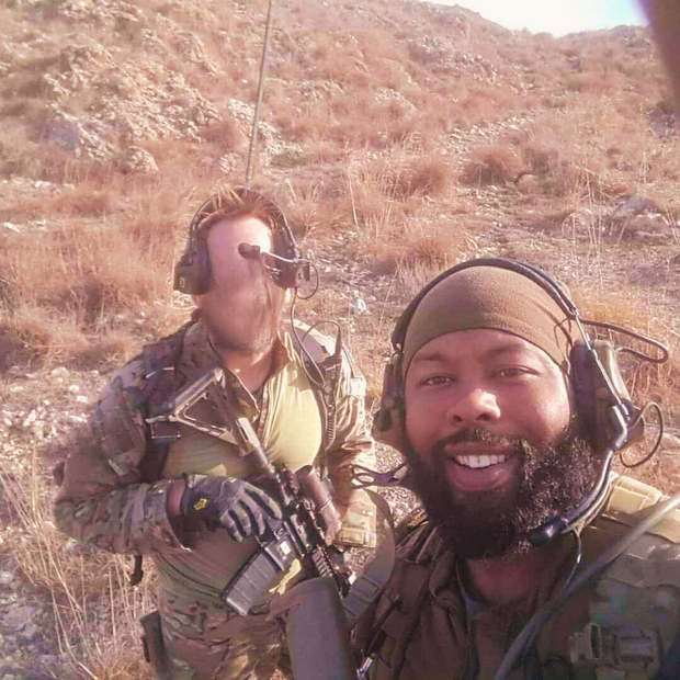 Two individuals in military gear with headphones in a desert-like environment