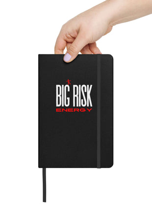 Person holding a Big Risk Energy notebook featuring a bold black cover and elastic closure, perfect for visionaries.