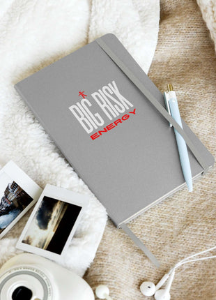 Big Risk Energy notebook on soft surface with photos and pen, ideal for visionaries and bold ideas.